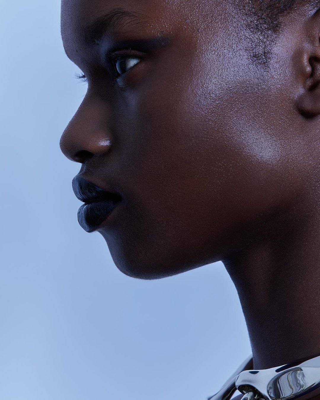 Close-up profile portrait of a Black woman with dark lipstick, wearing a metallic chain necklace against a light blue background.