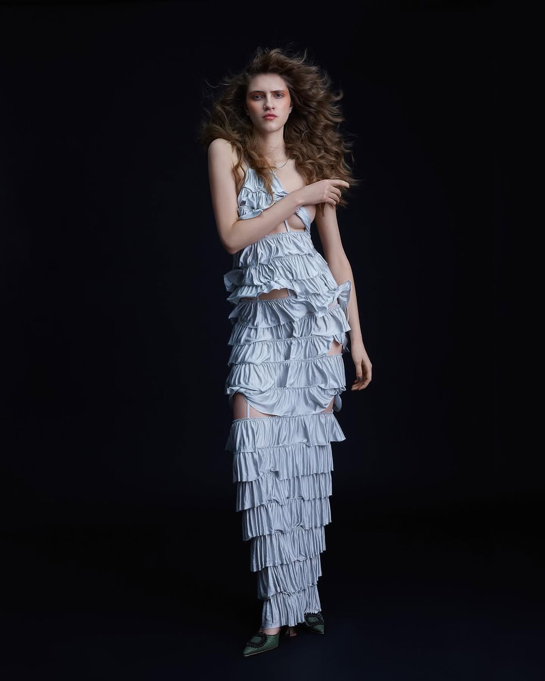 Fashion portrait of a woman in a layered ruffle dress with long curly hair posing against a dark background in a full shot.