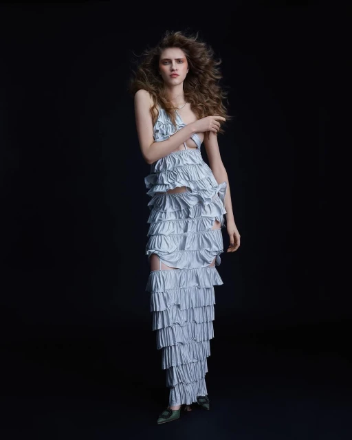 Fashion portrait of a woman in a layered ruffle dress with long curly hair posing against a dark background in a full shot.