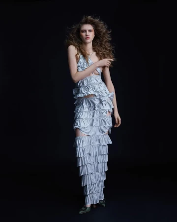 Fashion portrait of a woman in a layered ruffle dress with long curly hair posing against a dark background in a full shot.