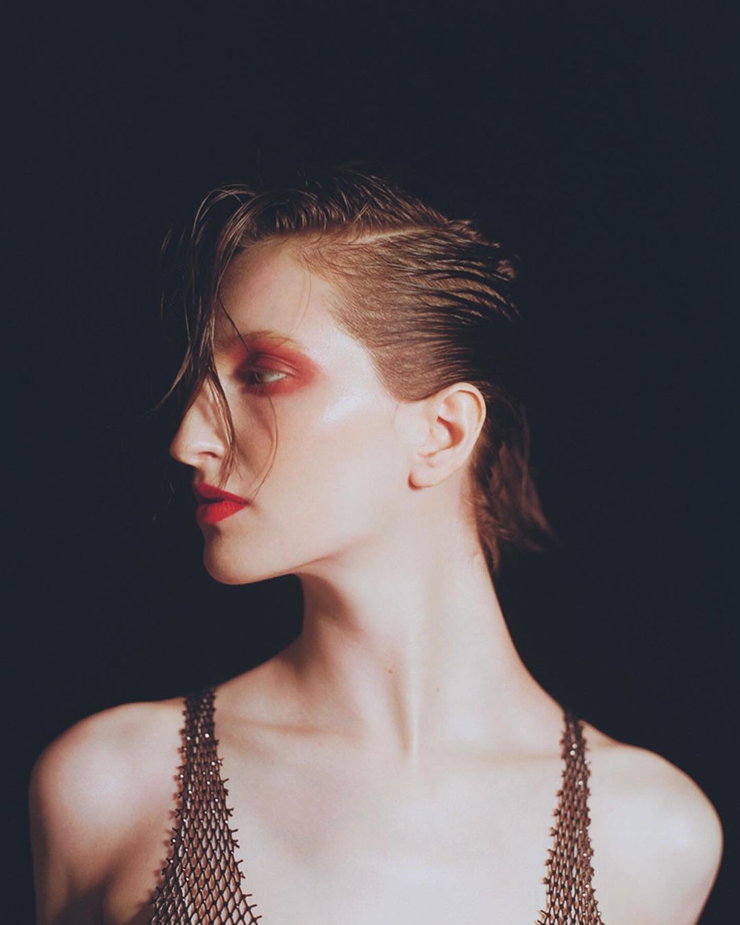 Elegant portrait of an adult woman with red makeup, an asymmetrical haircut and a chain-mail top against a dark backdrop in a studio.