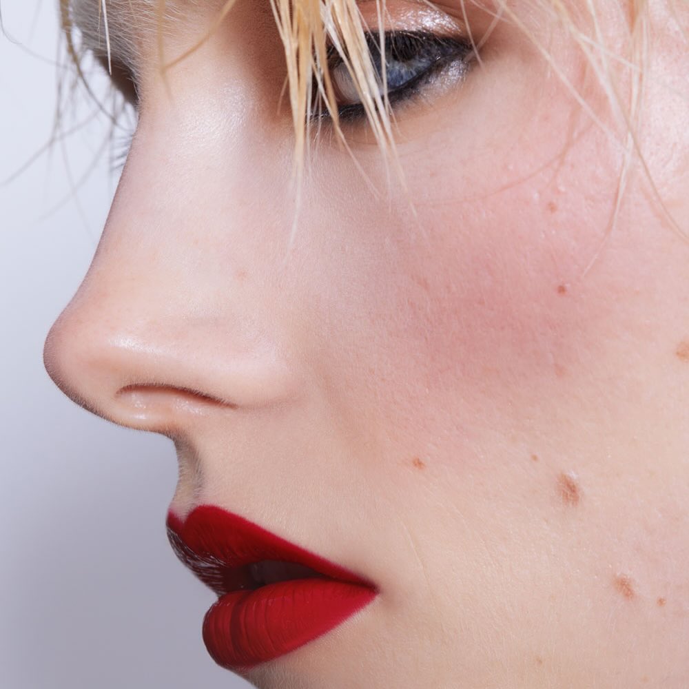 Close-up of a woman's face in profile. Visible are bright red lips, fair skin, blond hair, and blue eyes. The image is brightly lit.