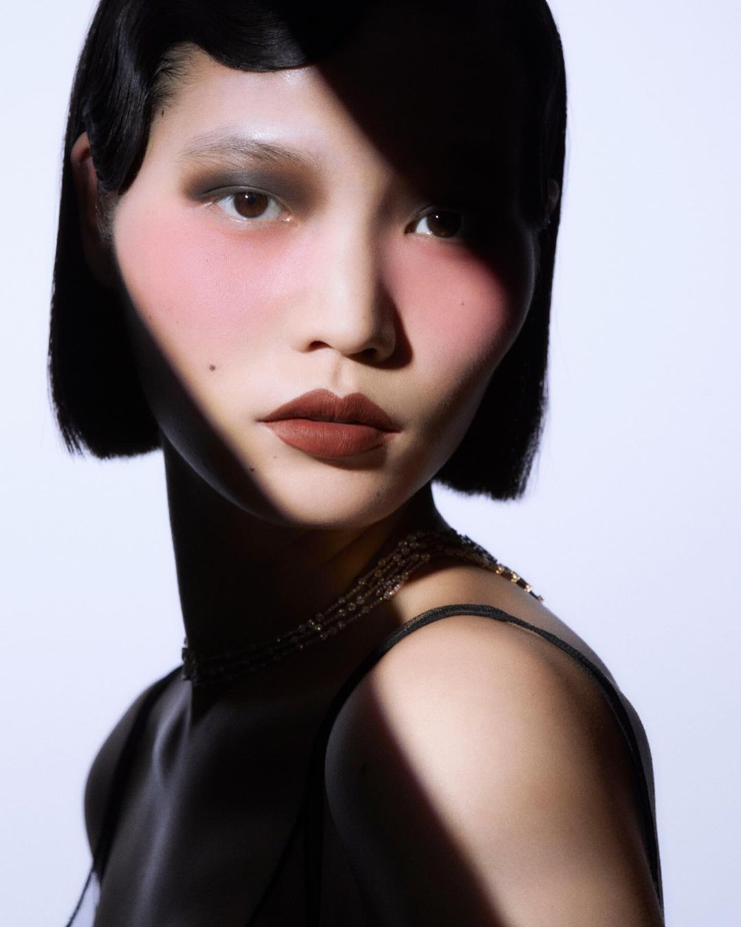 Close-up portrait of an Asian woman with short dark hair, wearing a gold necklace, and posing against a white background with shadow.
