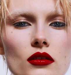 Close-up portrait of a woman with wet blonde hair, bleached eyebrows, dark blue eyes, glossy red lips, and pale skin.