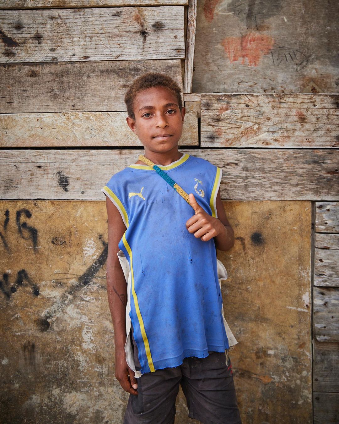 Young boy with curly hair in blue shirt, stands outdoors against weathered wooden and concrete wall, giving a thumbs up gesture.