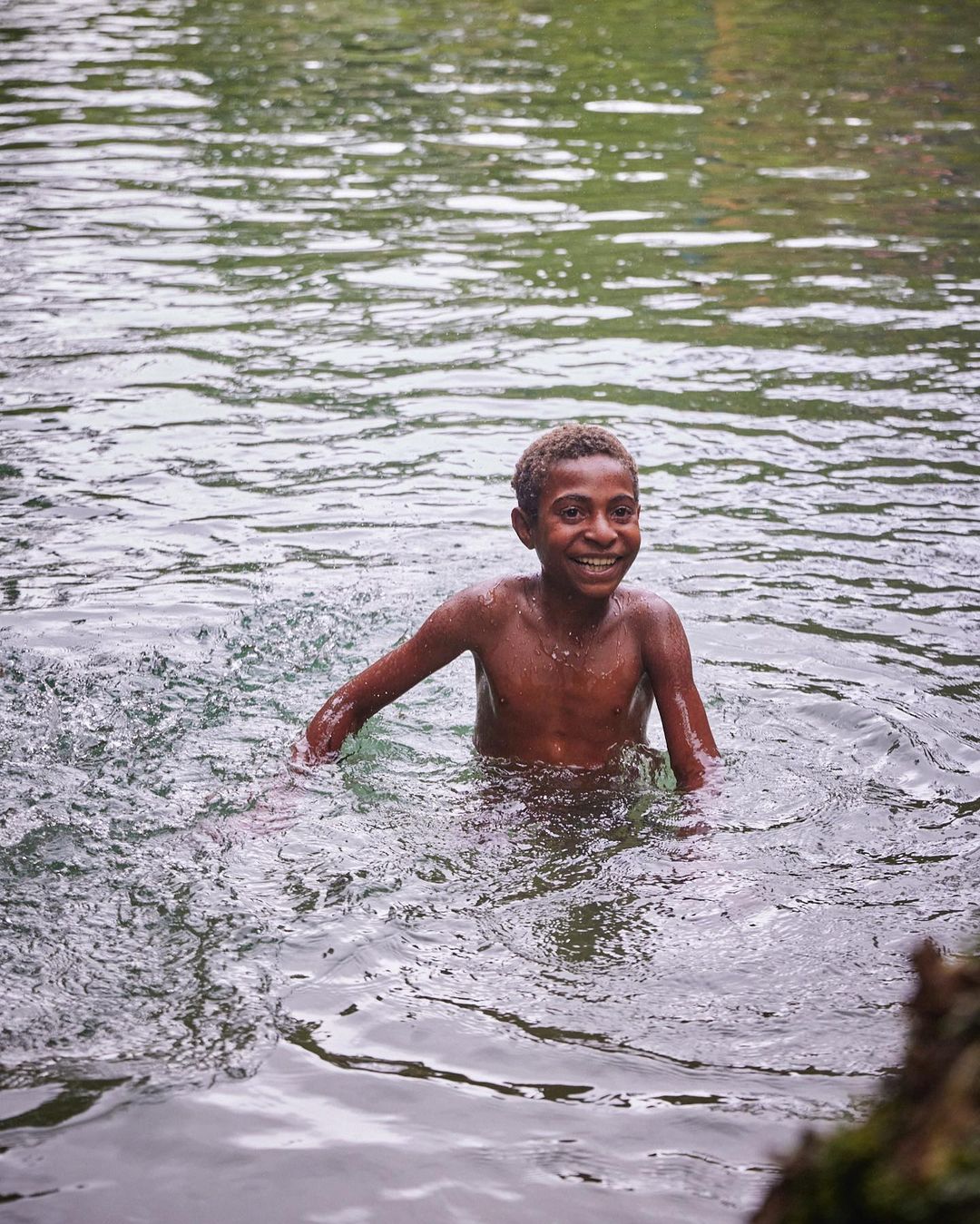 Young boy enjoys swimming in a river surrounded by lush greenery, smiling happily in a nature scene, showcasing an active lifestyle.