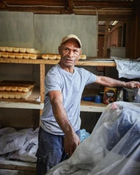 Man wearing a grey t-shirt and cap working inside a bakery with wooden shelves stacked with fresh bread