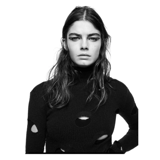 Monochrome portrait of a young adult female model with dark hair, wearing a black turtleneck sweater with cutouts, against a white backdrop.