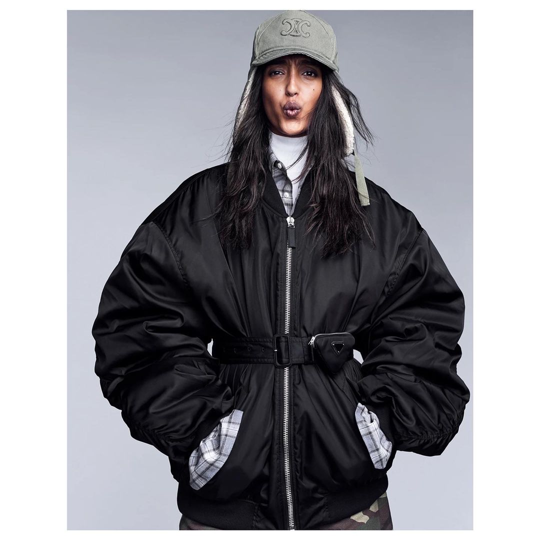 Stylish adult female posing in black oversized bomber jacket with Celine cap and Prada pouch on a neutral background in a studio shot.