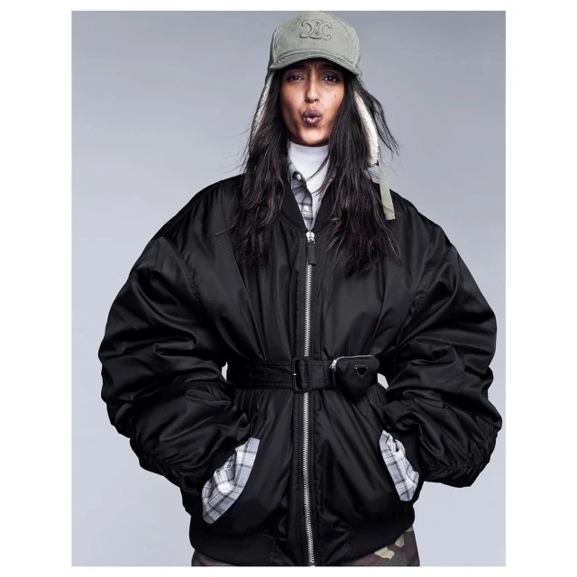 Stylish adult female posing in black oversized bomber jacket with Celine cap and Prada pouch on a neutral background in a studio shot.