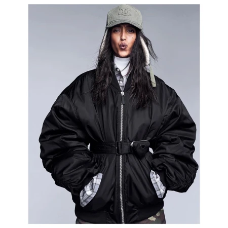 Stylish adult female posing in black oversized bomber jacket with Celine cap and Prada pouch on a neutral background in a studio shot.
