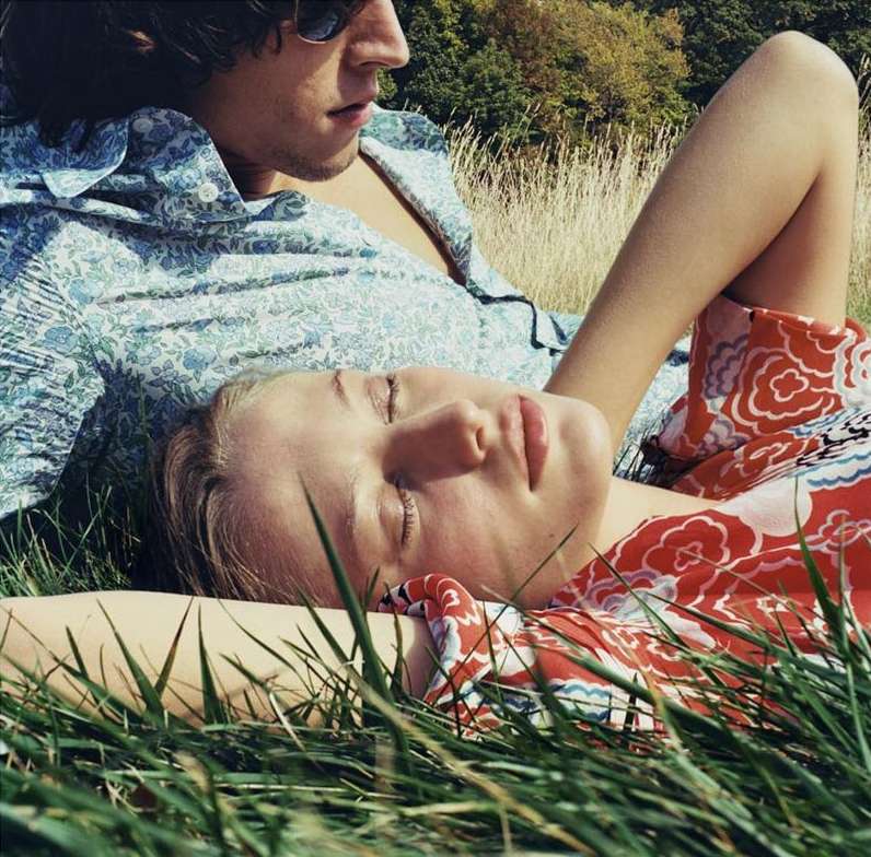 Woman rests in grassy field, wearing floral dress, eyes closed. Man with sunglasses lies behind her in blue patterned shirt, outdoors.