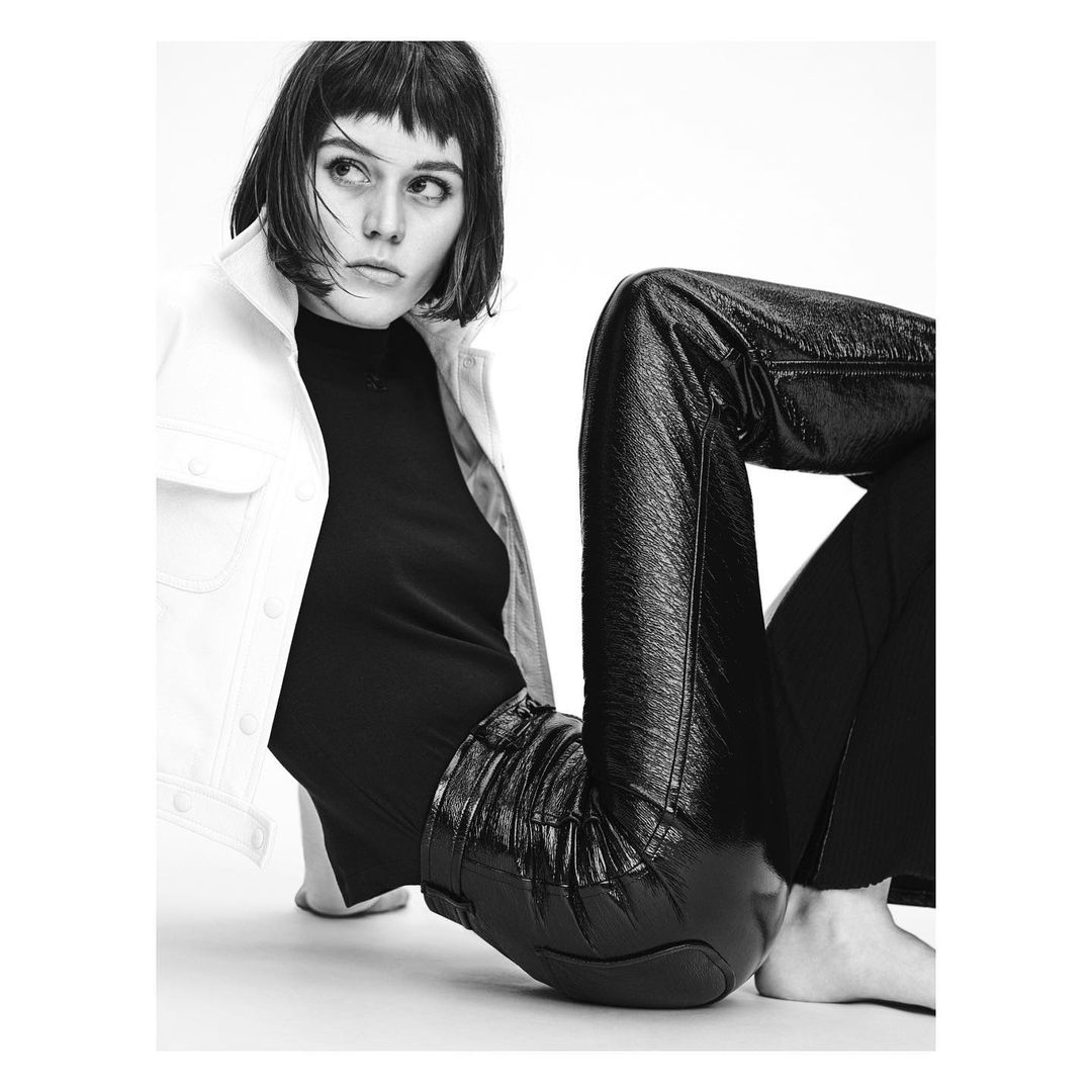 A stylish woman with a blunt haircut, wearing a white denim jacket, black top, and shiny black pants, poses against a white backdrop.