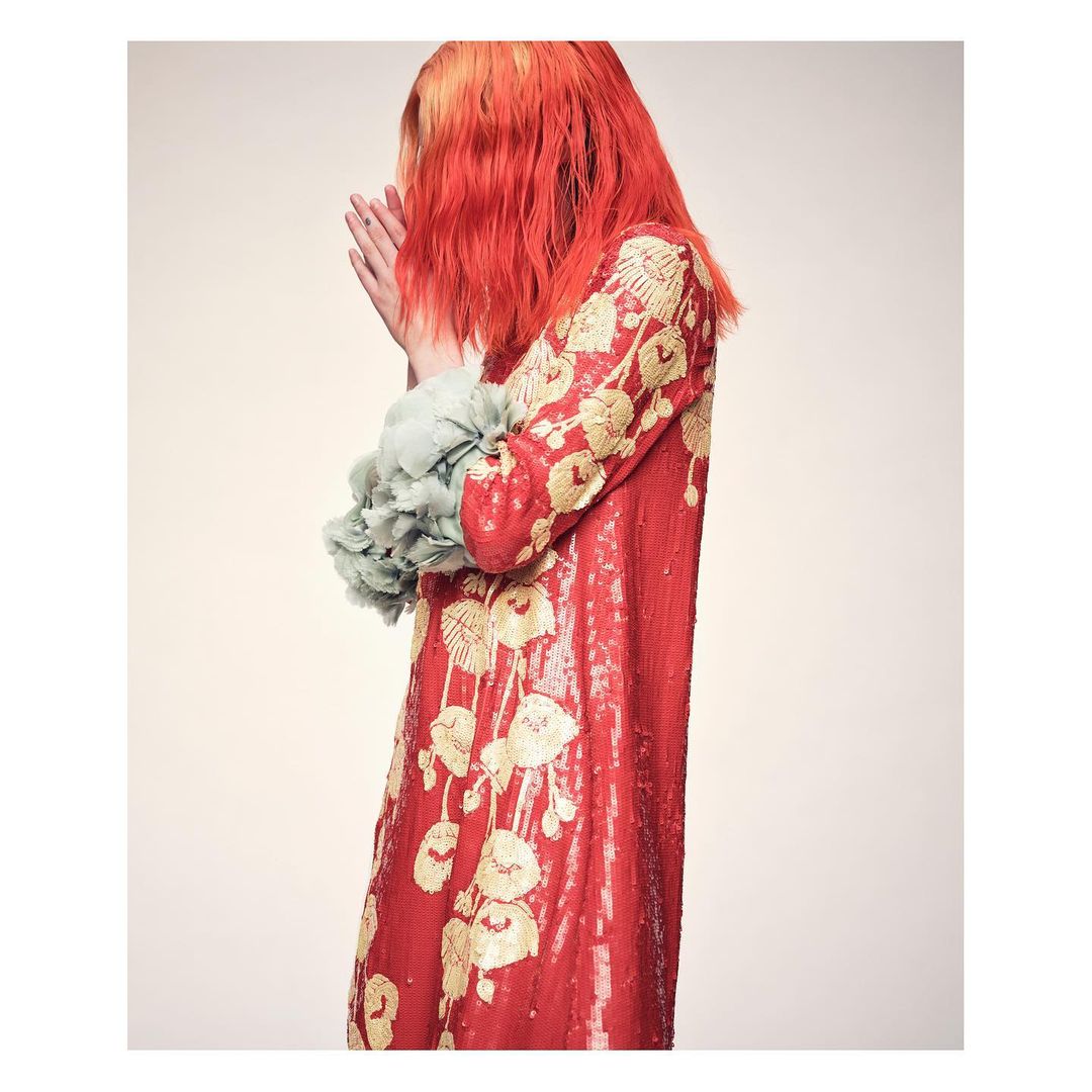 Red-haired person in a red sequin dress with floral embroidery and ruffled sleeves posing against a cream-colored background.