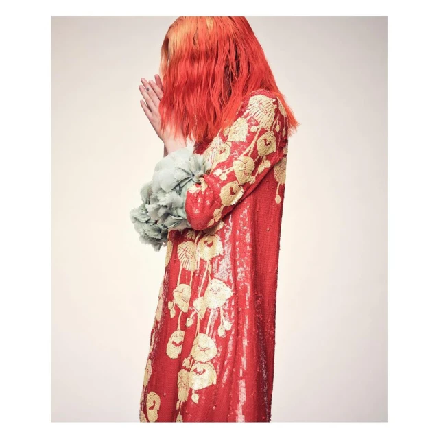 Red-haired person in a red sequin dress with floral embroidery and ruffled sleeves posing against a cream-colored background.