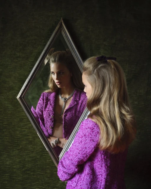 Blonde woman in purple jacket reflected in diamond-shaped mirror, against textured green backdrop, showcasing fashion and portraiture.