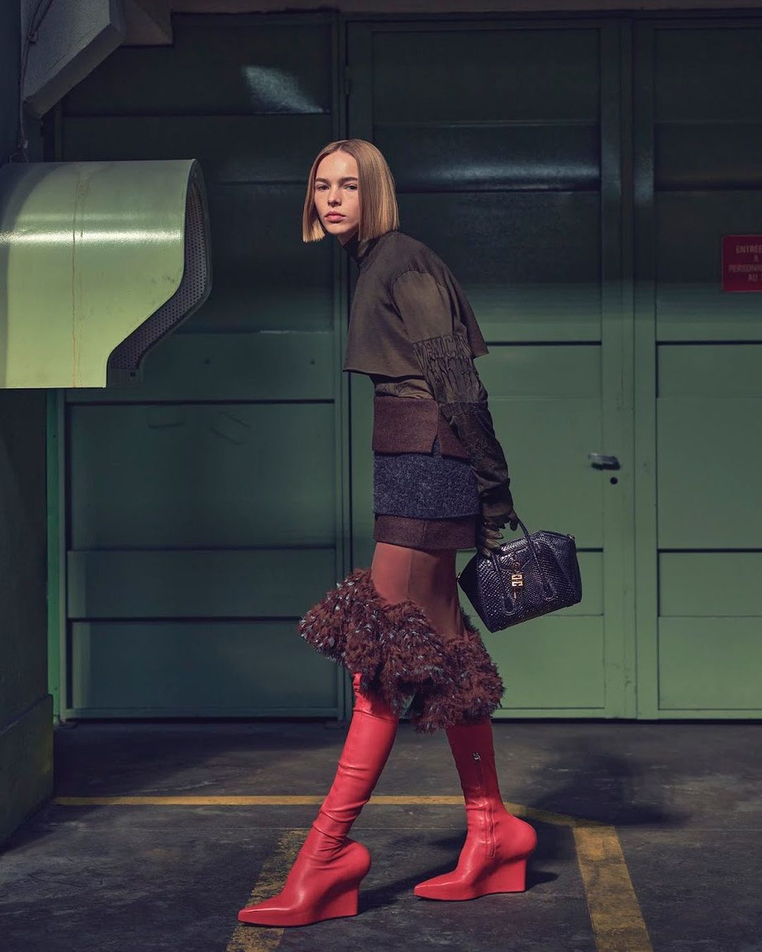 A woman in knee-high red boots and a layered outfit poses in a faded green industrial setting, holding a small black handbag.