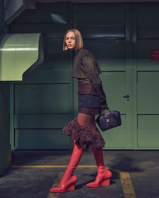 A woman in knee-high red boots and a layered outfit poses in a faded green industrial setting, holding a small black handbag.