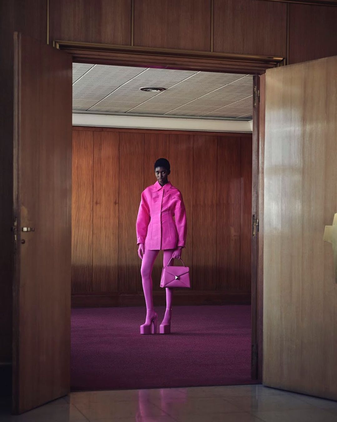 Fashion photo of an adult woman in a pink outfit with a pink purse and platform shoes standing in a room with wood walls and a door.