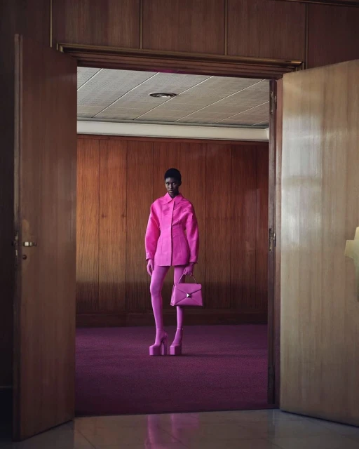 Fashion photo of an adult woman in a pink outfit with a pink purse and platform shoes standing in a room with wood walls and a door.
