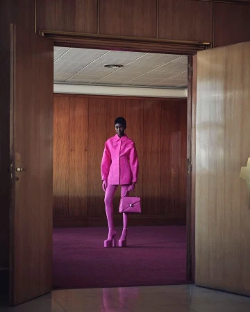 Fashion photo of an adult woman in a pink outfit with a pink purse and platform shoes standing in a room with wood walls and a door.