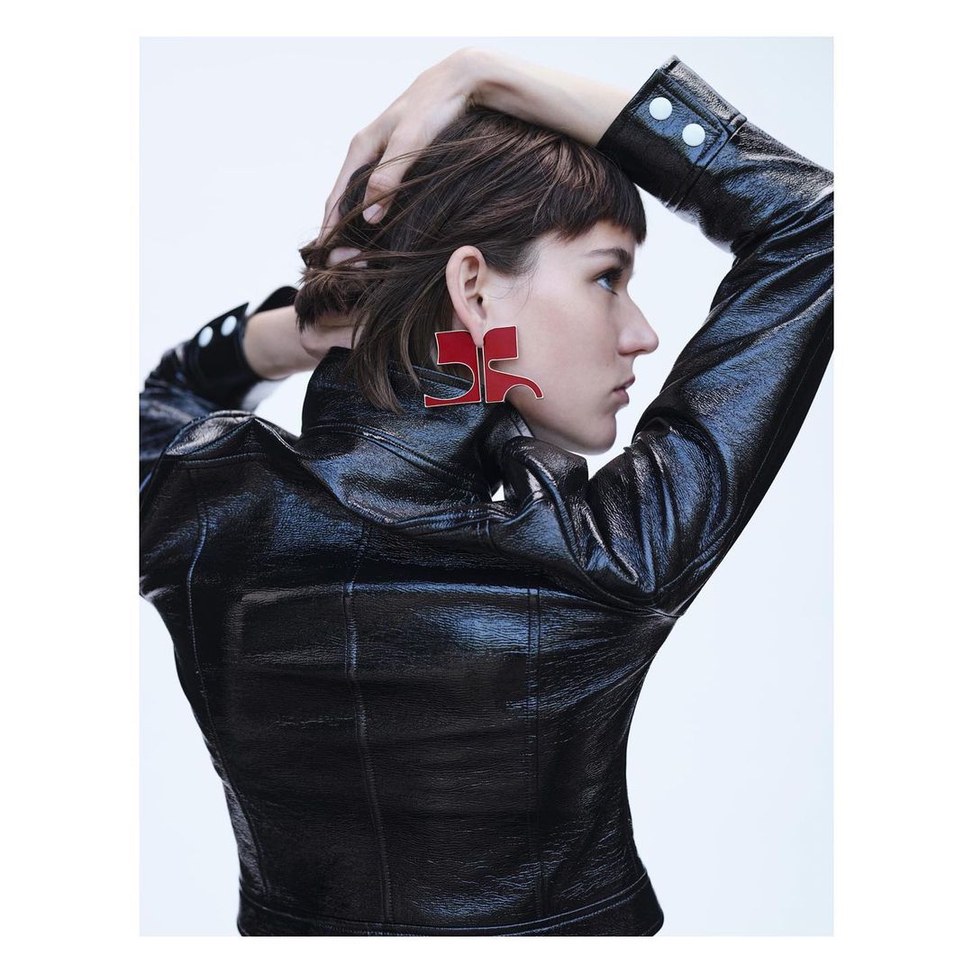 A profile view of a woman with dark hair wearing a black patent leather jacket and oversized red geometric earrings against a white background.