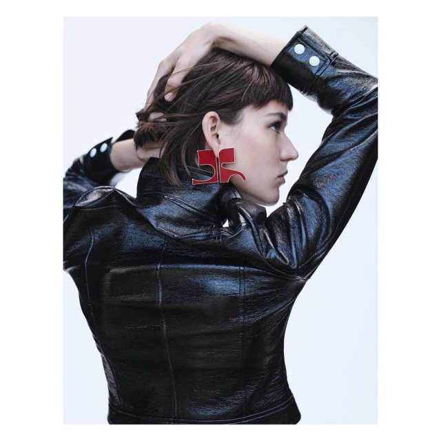 A profile view of a woman with dark hair wearing a black patent leather jacket and oversized red geometric earrings against a white background.