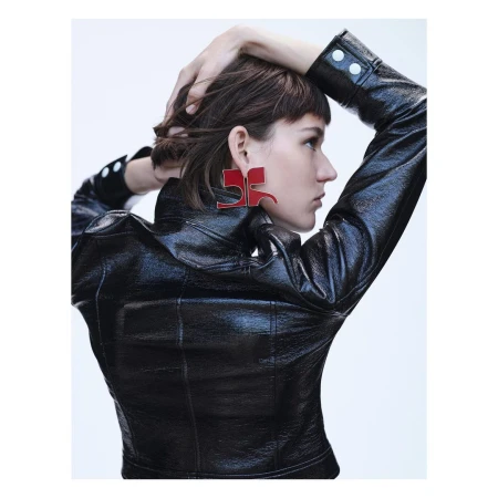 A profile view of a woman with dark hair wearing a black patent leather jacket and oversized red geometric earrings against a white background.