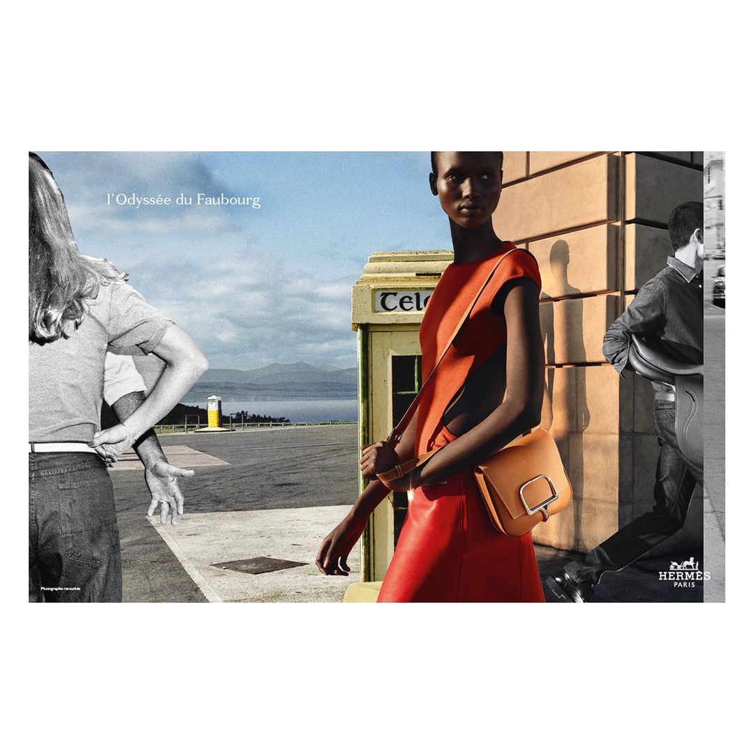 Fashion editorial shot of a black woman in an orange dress with a brown purse, set against an outdoor backdrop with black-and-white figures.
