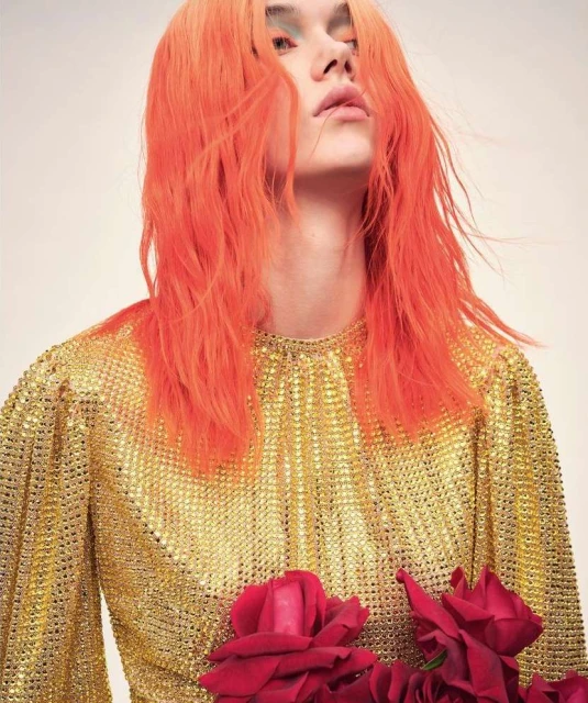 Fashion portrait of a young adult with orange hair wearing a gold sequin blouse, holding red roses, posing in a photo studio.