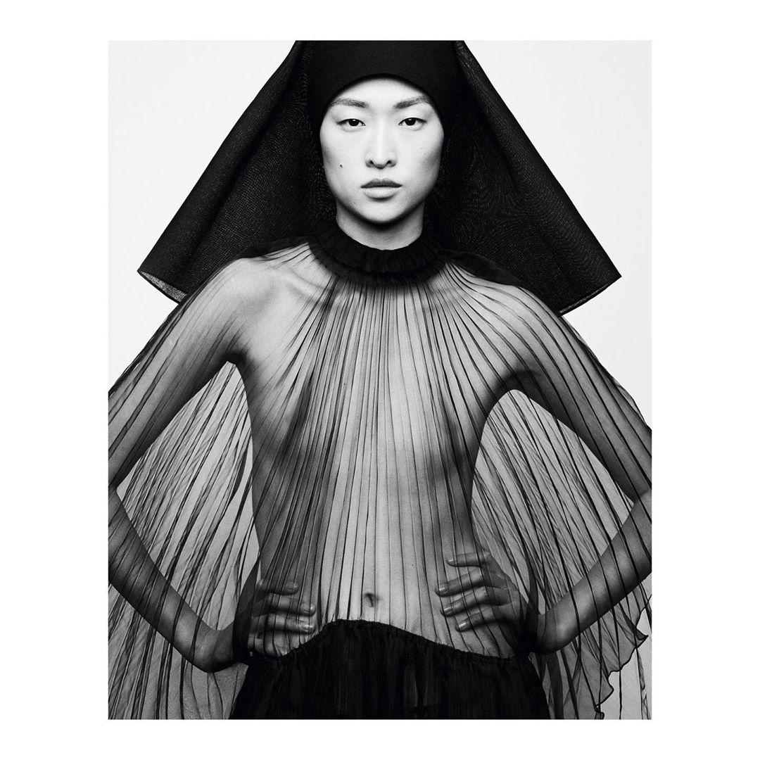 Asian woman in a black headpiece and sheer, pleated black dress poses against a white background in a studio shot.