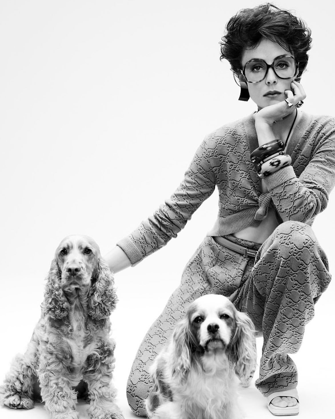 Black and white portrait of a woman with glasses kneeling with two dogs, wearing a patterned sweater and pants in a white studio.