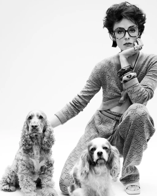 Black and white portrait of a woman with glasses kneeling with two dogs, wearing a patterned sweater and pants in a white studio.
