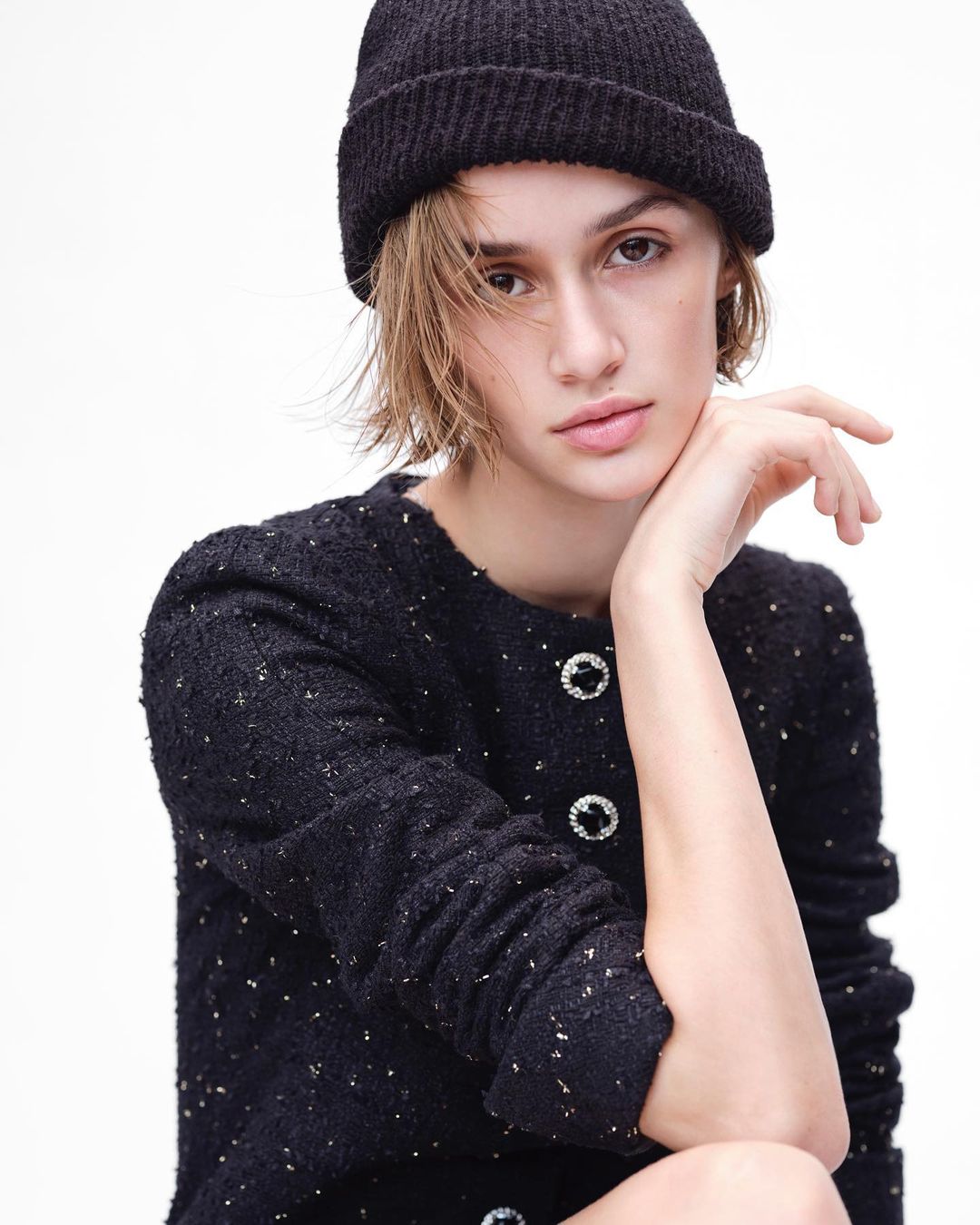 Close-up of a woman with light skin wearing a black beanie and black jacket with decorative buttons in a bright, neutral studio.