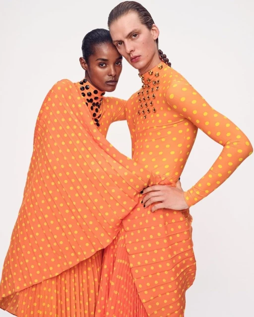 Two models pose indoors, wearing orange polka dot dresses with embellished necklines. One model has a draped cape sleeve.