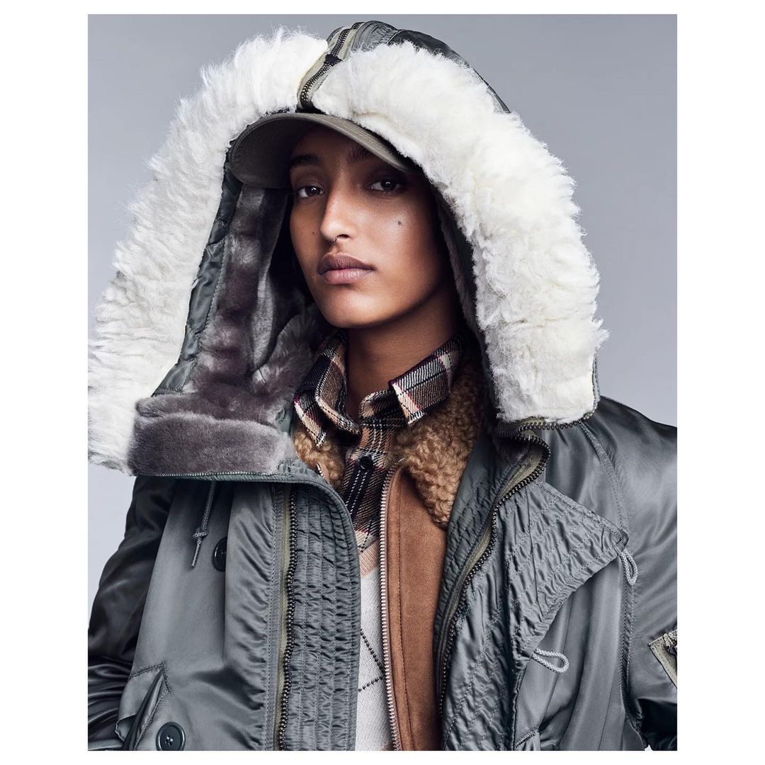Close-up portrait of a woman wearing an olive-green hooded parka with fur trim, posing in a studio with soft, neutral lighting.