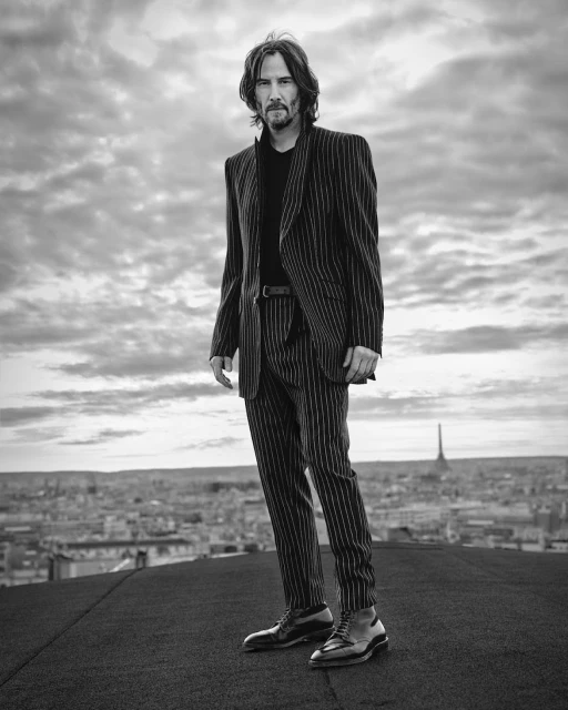 Black and white portrait of Keanu Reeves in a pinstripe suit standing on a rooftop in Paris with the Eiffel Tower visible in the background.