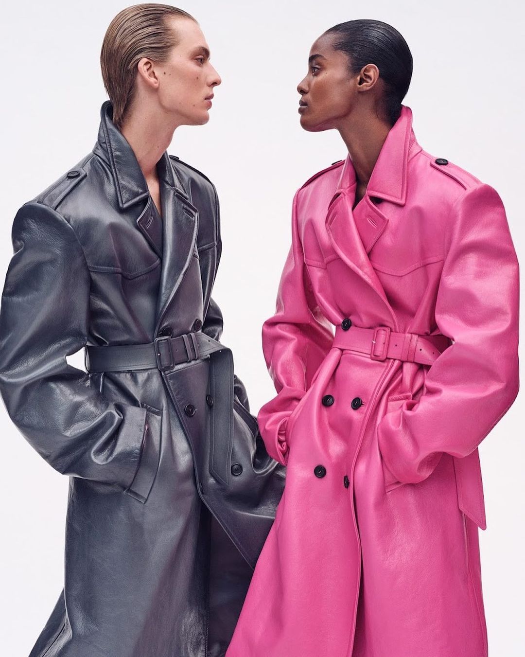 Two fashion models wearing full-length glossy trench coats in pink and grey, face each other in a fashion forward studio shot.