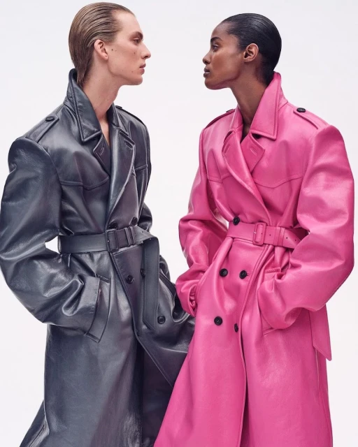 Two fashion models wearing full-length glossy trench coats in pink and grey, face each other in a fashion forward studio shot.