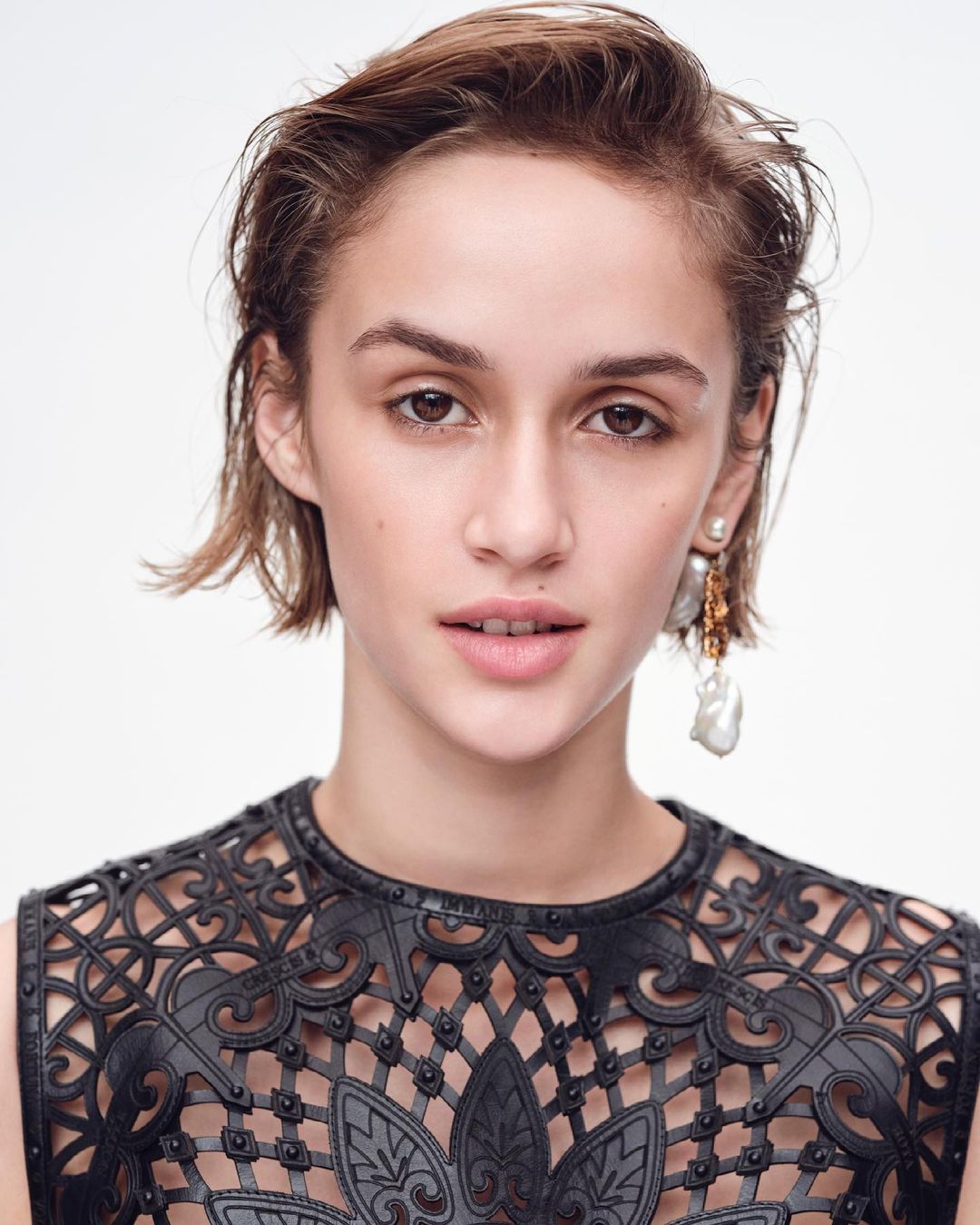 Close-up portrait of a young woman with short brown hair, wearing a black patterned top and pearl earrings, against a white background.