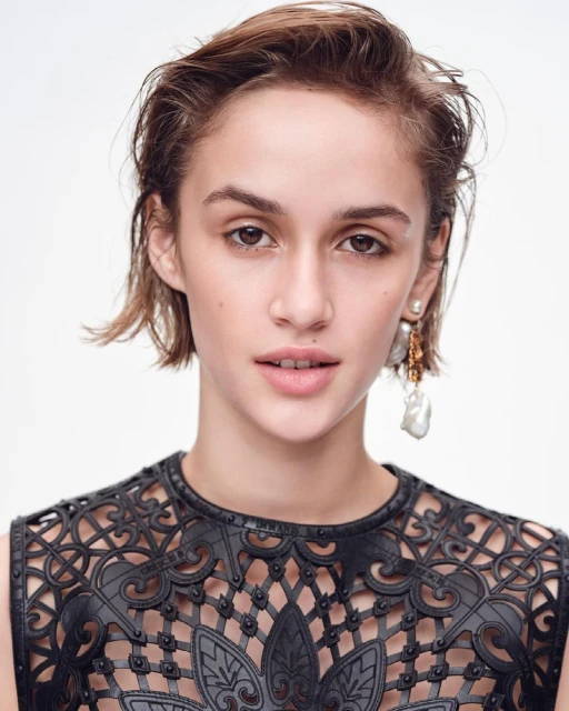 Close-up portrait of a young woman with short brown hair, wearing a black patterned top and pearl earrings, against a white background.