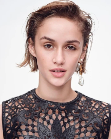 Close-up portrait of a young woman with short brown hair, wearing a black patterned top and pearl earrings, against a white background.
