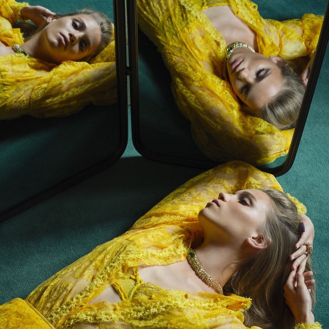 Woman in yellow lace dress lies on a green surface, reflected in a black-framed mirror. Jewelry adorns her neck and fingers.