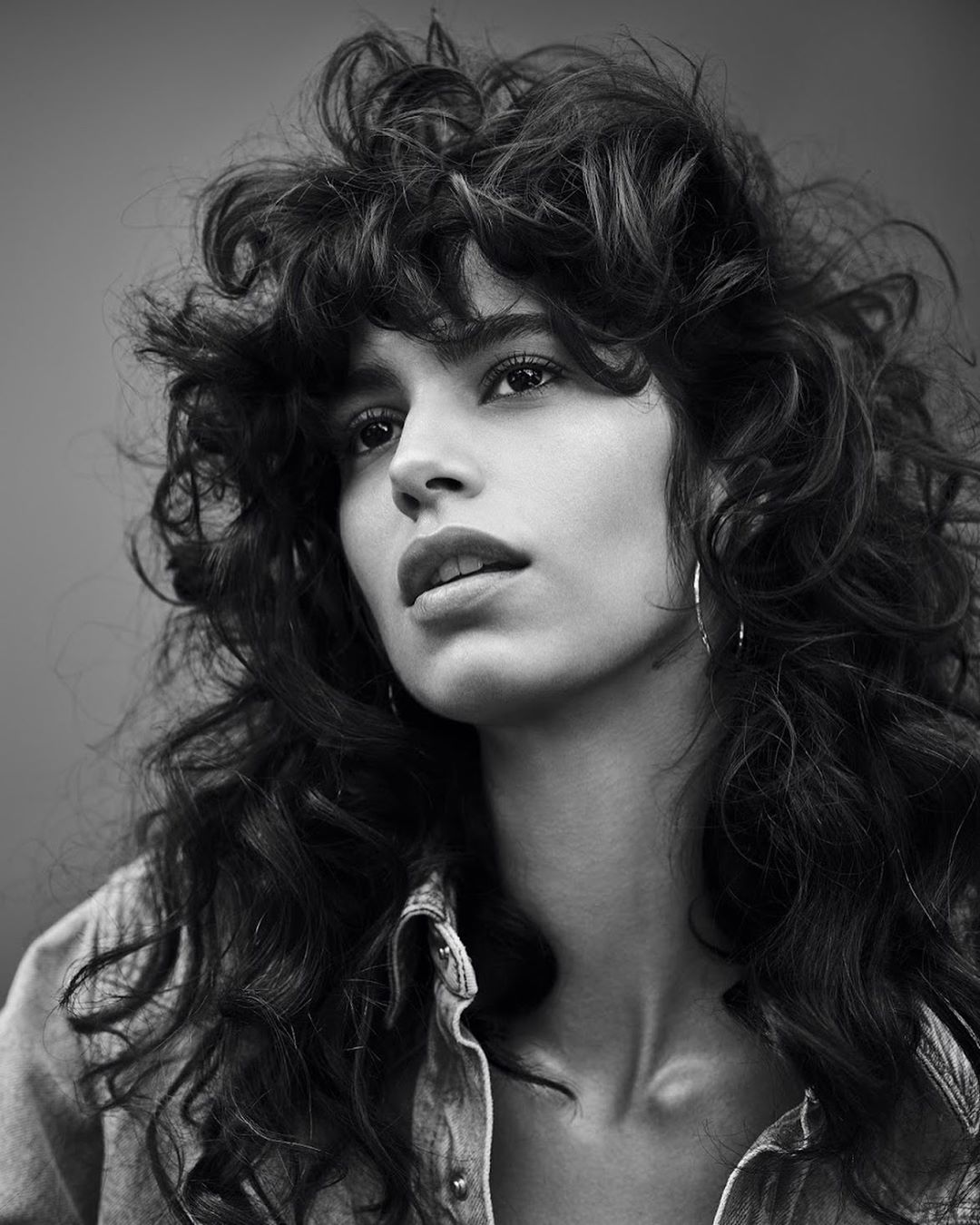 Black-and-white portrait of a woman with curly hair, wearing a denim jacket, and hoop earrings against a neutral backdrop.