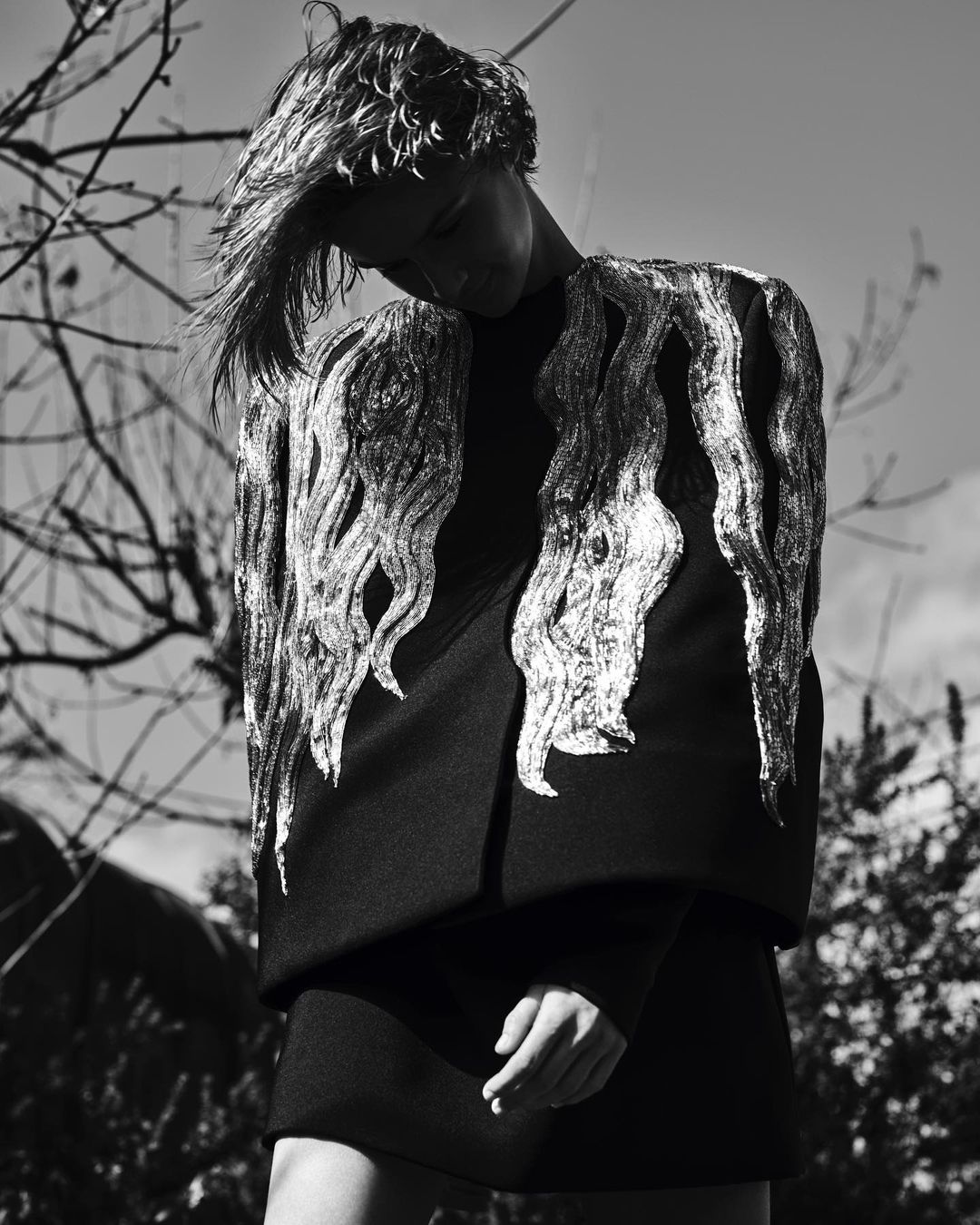 Black and white fashion shot of an adult woman with blonde hair wearing a black dress with silver shoulder embellishments outdoors.