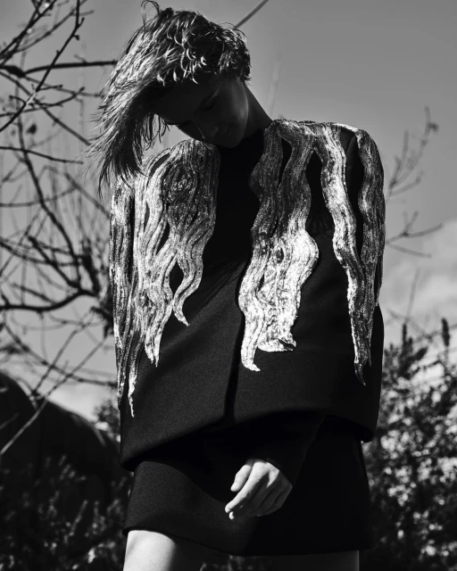 Black and white fashion shot of an adult woman with blonde hair wearing a black dress with silver shoulder embellishments outdoors.