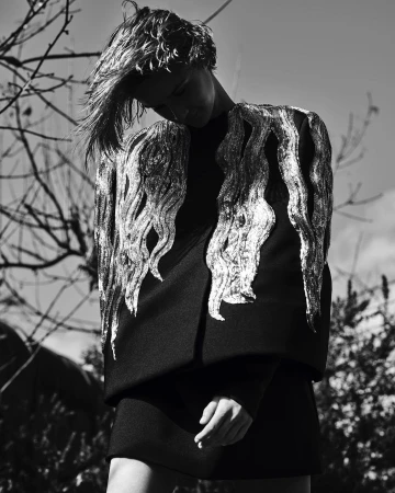 Black and white fashion shot of an adult woman with blonde hair wearing a black dress with silver shoulder embellishments outdoors.