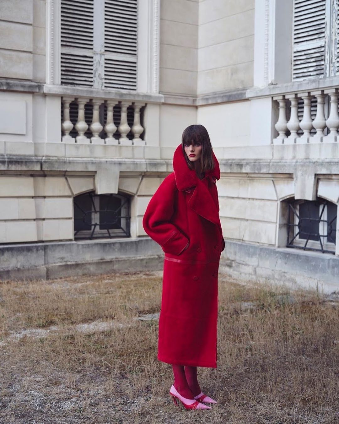 Fashionable woman in a red overcoat and pink heels stands outside a building with closed shutters in a style shot, creating a visual harmony.