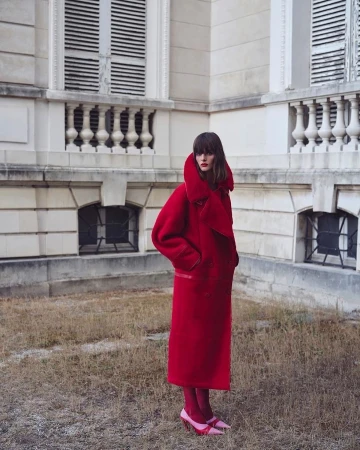 Fashionable woman in a red overcoat and pink heels stands outside a building with closed shutters in a style shot, creating a visual harmony.