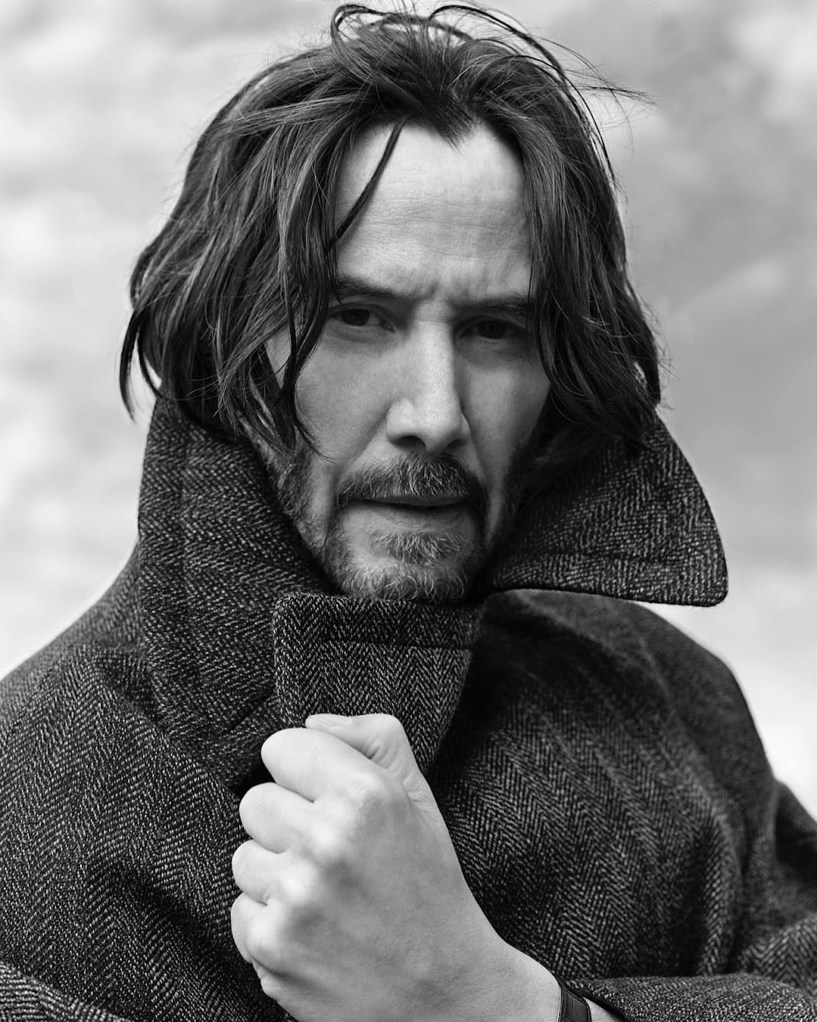 Black and white portrait of Keanu Reeves wearing a herringbone coat, looking at the camera with a neutral expression, against a blurred background.